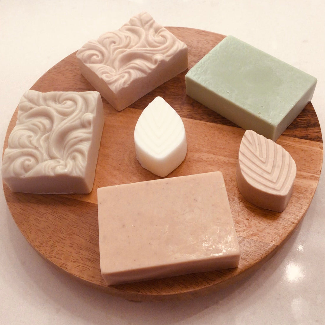 Assorted handcrafted clay soap bars arranged on a round wooden board, including French Green, Kaolin, and Red Moroccan clay bars by Handcrafted Soaps by SynEb