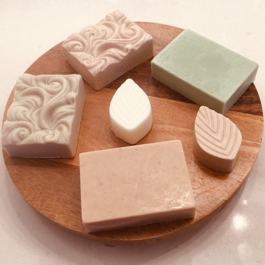 Assorted handcrafted clay soap bars arranged on a round wooden board, including French Green, Kaolin, and Red Moroccan clay bars by Handcrafted Soaps by SynEb